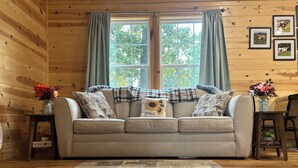 A cozy room with a wooden interior, a beige sofa with patterned cushions, a window with curtains, and a rug on the floor. - Texas Longhorn Ranch-Private Cabin-Pasture Views (Licking)