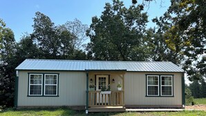 A small, single-story house with a porch and a metal roof, surrounded by grass and trees. - Texas Longhorn Ranch-Private Cabin-Pasture Views (Licking)