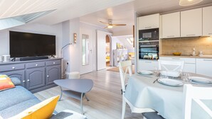 A modern living space with a flat-screen TV, grey cabinets, a dining area with a table set, and a kitchen with a microwave. - Apartment 'Les Hauts Du Lac' with Lake View and Wi-Fi (Les Sables-d'Olonne)