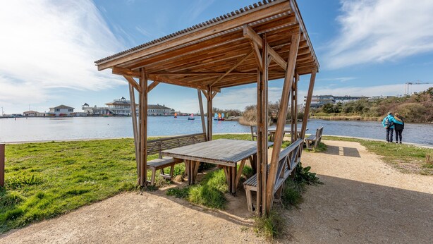 A wooden pavilion with a table and benches, overlooking a body of water with boats and buildings in the background. - Apartment 'Les Hauts Du Lac' with Lake View and Wi-Fi (Les Sables-d'Olonne)