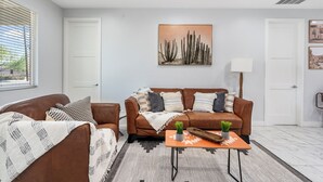 A modern living room with a brown leather sofa, a matching armchair, and a coffee table with decorative items. A large painting of cacti hangs on the wall, and there is a window with blinds on the left.