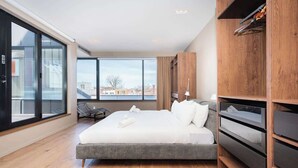 A modern hotel room with a large bed, wooden flooring, a view through floor-to-ceiling windows, and built-in storage units. - Luxury 3+ BR Penthouse Near Jean-Talon Market (Montréal)