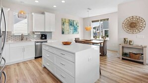 A modern kitchen with white cabinetry, a central island, and a dining area with a wooden table and chairs. - Family-Friendly Home with Rooftop Lounge (Inlet Beach)