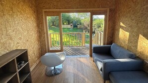 A room with a wooden ceiling, a sliding glass door leading to a balcony with a wooden railing, a grey sofa, and a modern chair.