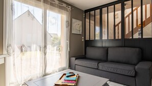 A room with a grey sofa, a white table with a tray of books, sheer curtains with decorative patterns, and a staircase with wooden railings. - Chez Patricia et Patrice by Interhome (Guissény)