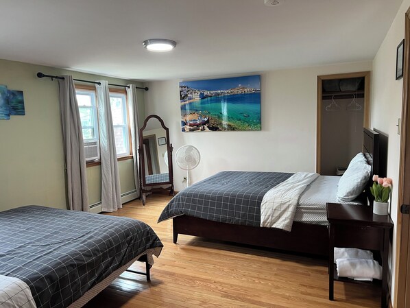 A bedroom with two beds, a large window with curtains, a mirror, a painting of a coastal scene, and a wooden floor.