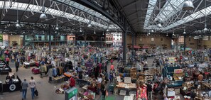 Explore a vibrant marketplace with numerous stalls and vendors, set beneath a high ceiling with metal beams and large lamps.