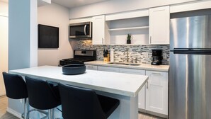 Une cuisine moderne avec des armoires blanches, un réfrigérateur en acier inoxydable, un four à micro-ondes et une machine à café. On y trouve un petit îlot central avec un récipient et des chaises noires.
