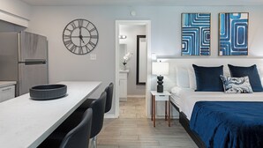 Une chambre d’hôtel moderne dotée d’un coin cuisine, d’un lit avec une literie bleue, d’une grande horloge murale et d’œuvres d’art abstraites accrochées au mur.