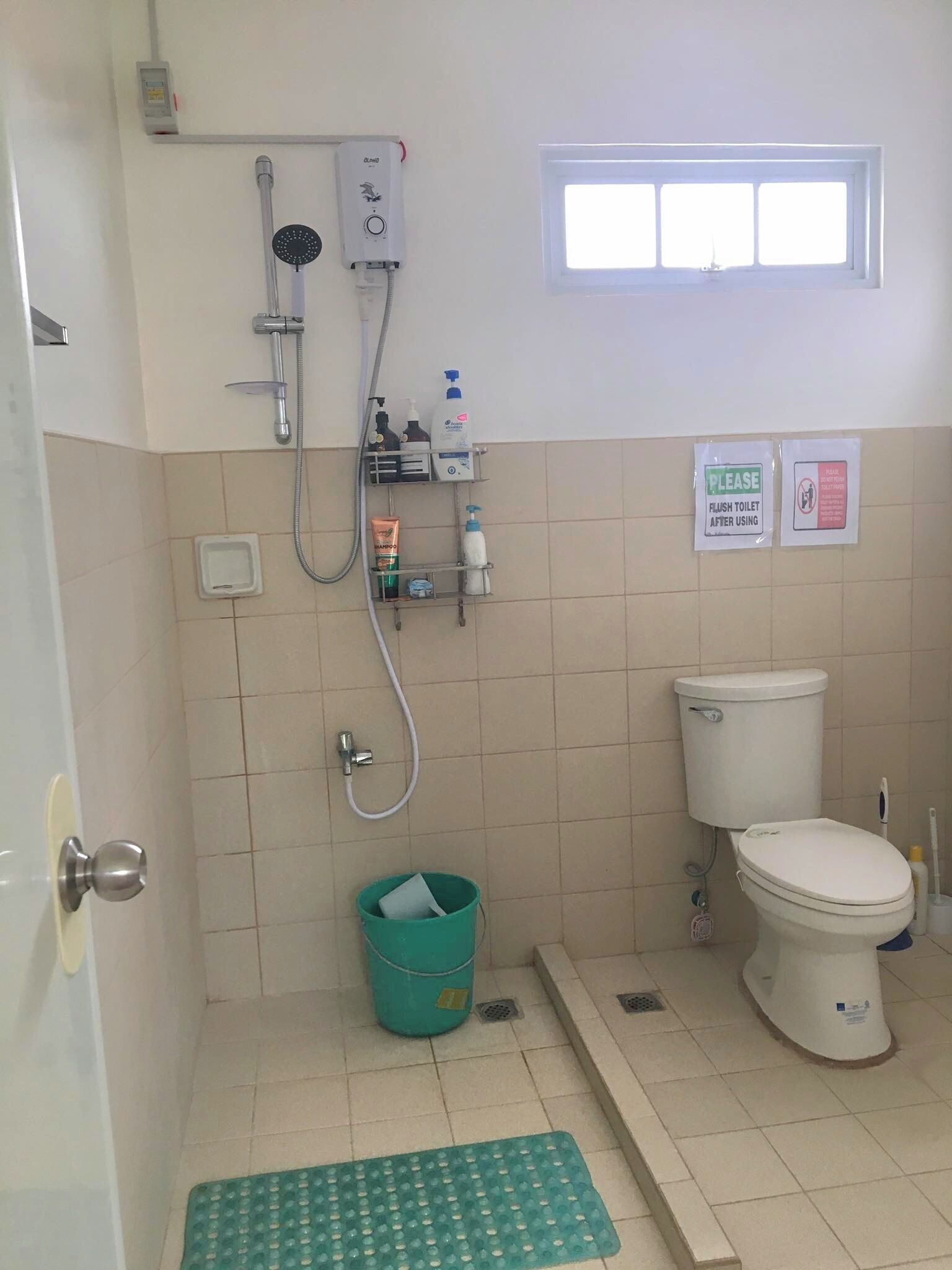 A small bathroom with a toilet, a shower, and a window. There are toiletries on a shelf and a sign instructing to flush the toilet after use.