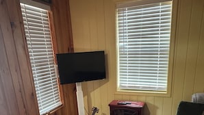 A cozy room with wooden walls, a flat-screen TV mounted on the wall, a guitar, a small wooden table, a tufted ottoman, and a colorful rug. - Cozy cabin in pleasant Brewton with AC, WiFi, and fire pit.  (Brewton)