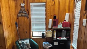 A small cabin with wooden walls, a teal chair, a small kitchen area with a refrigerator, microwave, and coffee maker, and a window with blinds. - Cozy cabin in pleasant Brewton with AC, WiFi, and fire pit.  (Brewton)