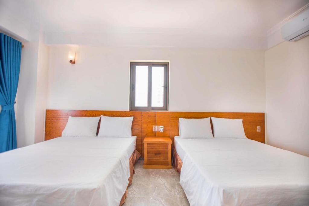 A hotel room with two single beds, a wooden headboard, a small bedside table, a window, and a wall-mounted air conditioning unit.