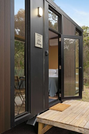 A modern, compact room with a bed, a small table, and a bench outside the door. - Bushland Haven by Tiny Away (Cashmore)