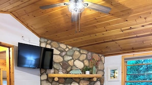 A cozy living room with a stone fireplace, wooden ceiling, and a television mounted on the wall.