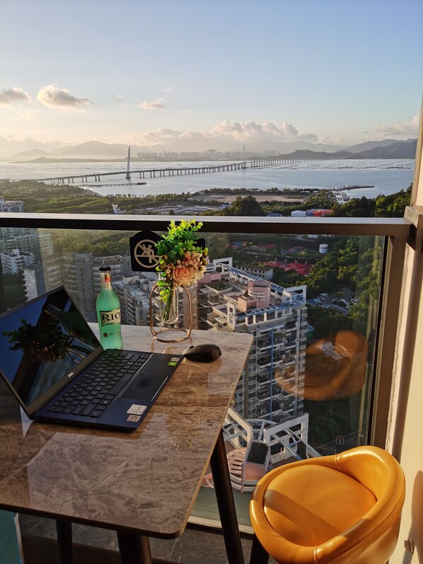 A laptop on a table with a bottle of RIO, a vase with green plants, and a mouse. A yellow chair is next to the table. A large window offers a view of a bridge and buildings across a body of water.