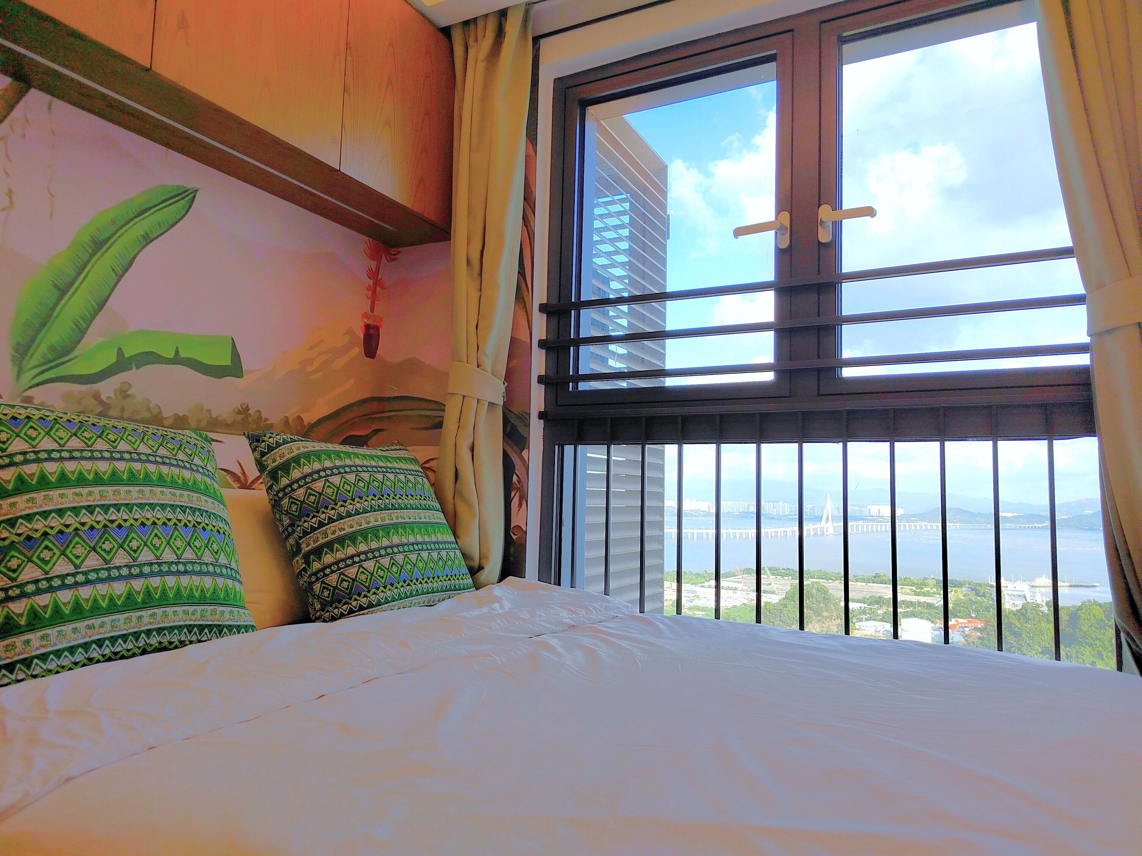 Habitación de hotel con cama, almohadones estampados, un mural y vistas a un puente y al agua a través de amplias ventanas.
