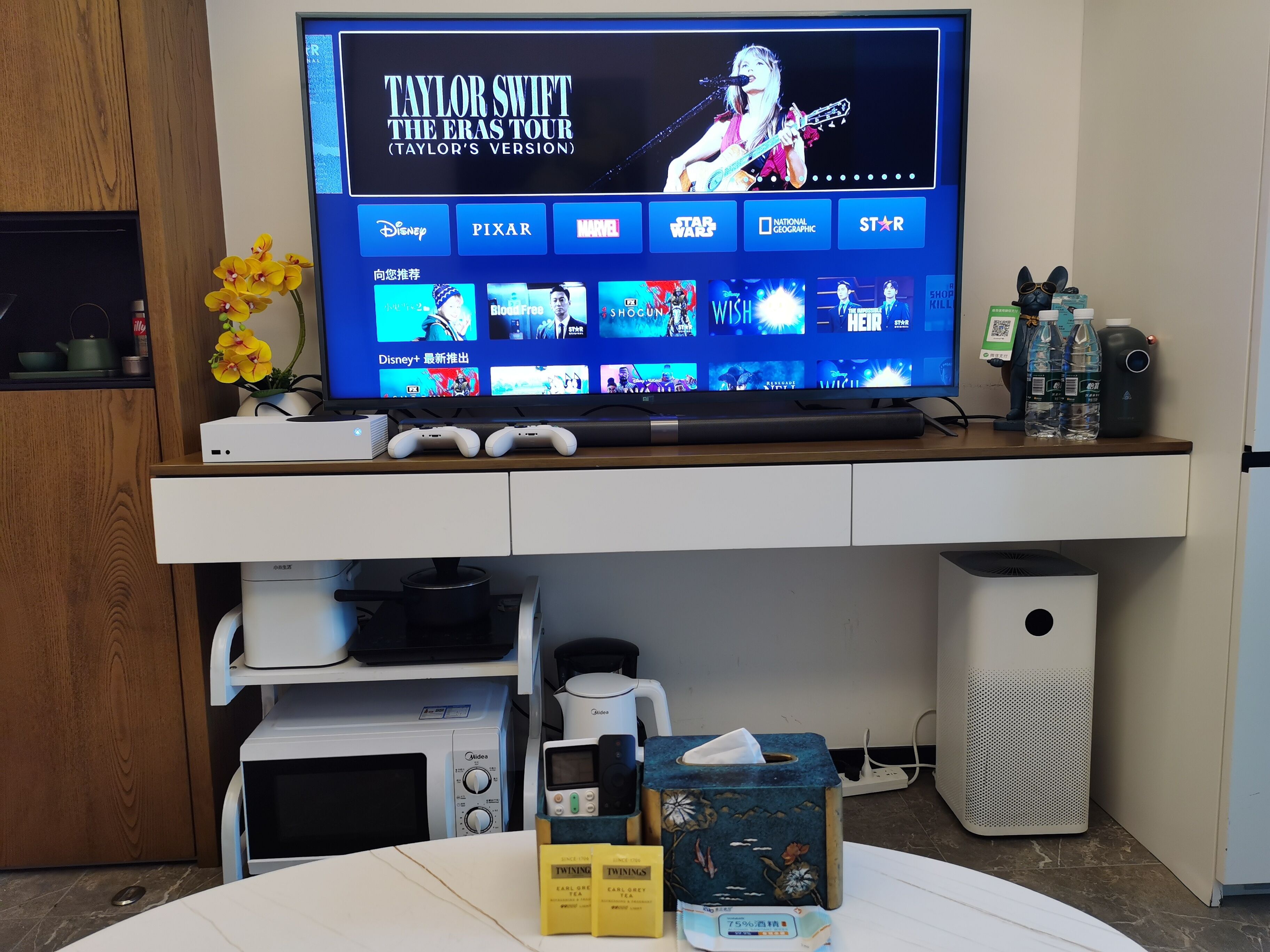 Una sala de estar moderna con televisor de pantalla plana mostrando la gira de un artista musical, una cafetera, un microondas y un altavoz.