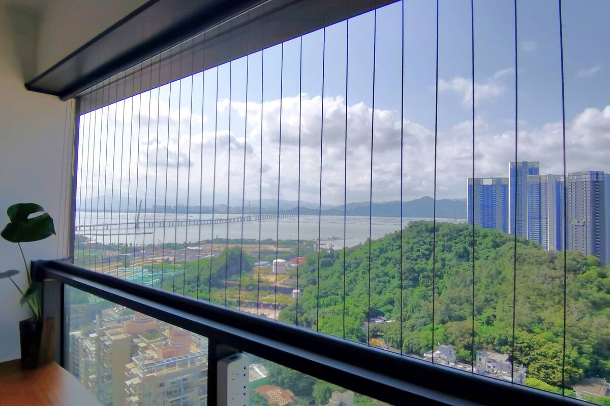 A view from a balcony with a metal railing, overlooking a river, green hills, and a bridge.