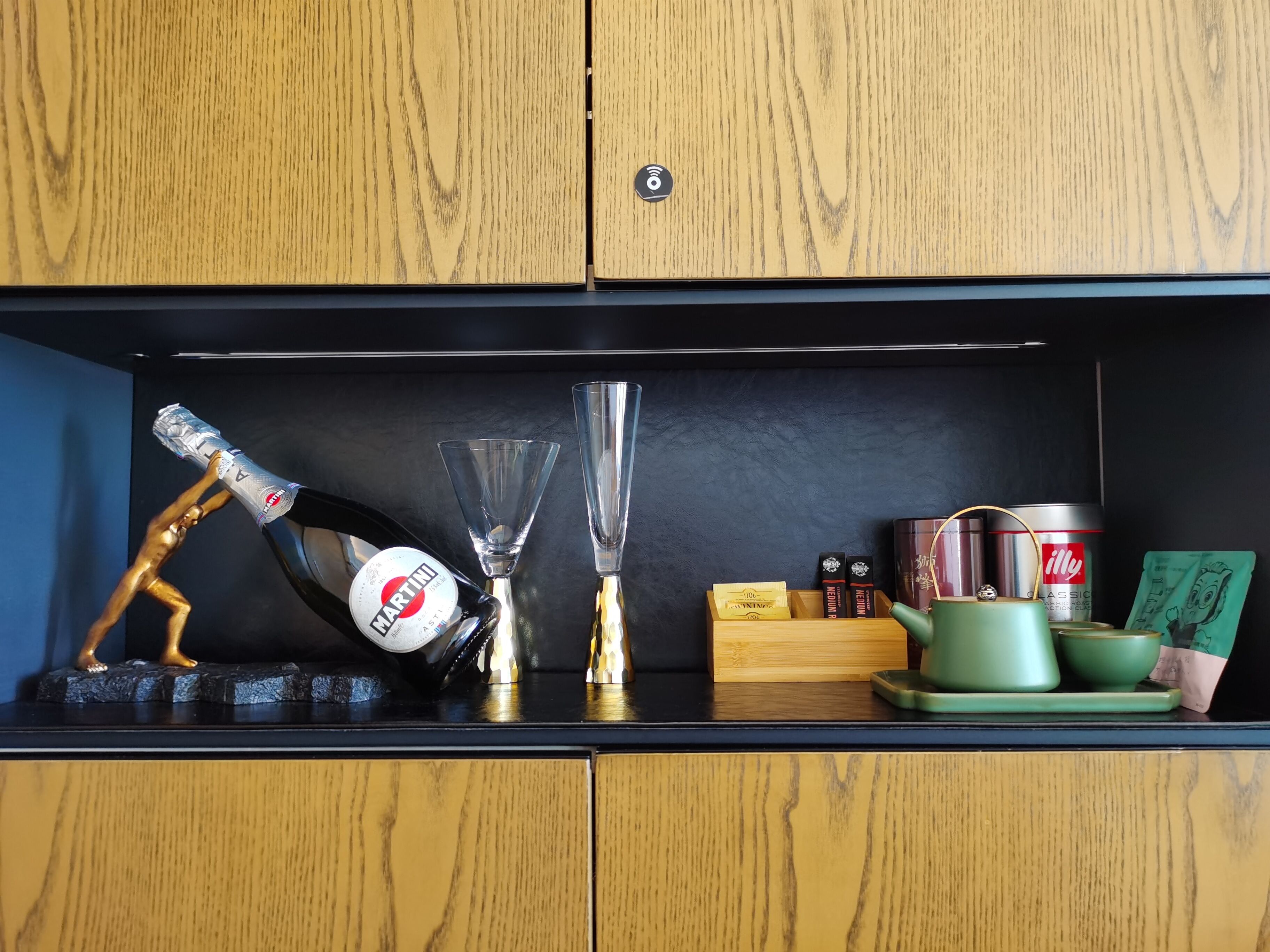 Un mueble de madera con una encimera negra. Sobre la encimera, hay una botella de Martini, dos cocteleras, una tetera verde, una taza de té verde y una caja de café Illy.