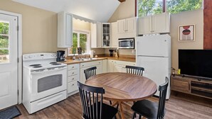 A kitchen with white cabinets, a stove, a microwave, and a dining table with chairs. - Forest cabin w/ hot tub, deck & huge yard (Gerrardstown)
