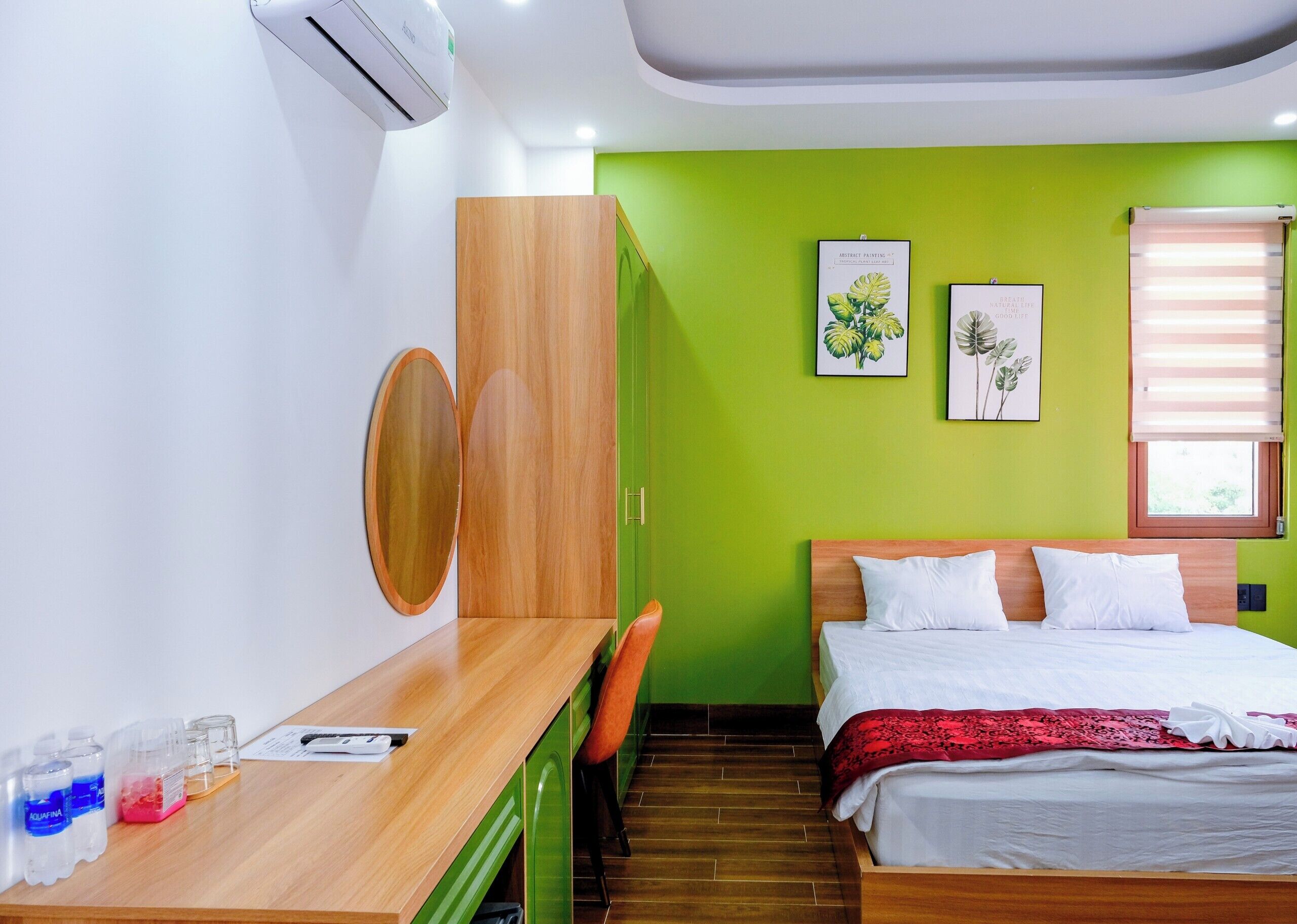 A modern hotel room with a wooden desk, a bed with white bedding, green walls, and two framed botanical prints.