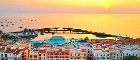 A coastal cityscape with colorful buildings, a prominent bridge, and a calm sea at sunset.