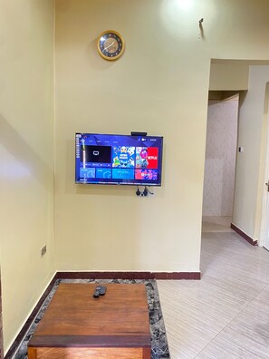 A living room with a mounted flat-screen TV displaying various apps and a clock on the wall. - Spacious 2-Bedroom Apartment in Zanzibar with Free Parking & Entertainment (Zanzibar)