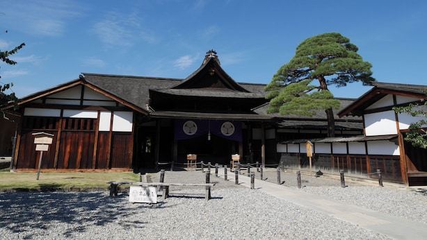 Edificio japonés tradicional con estructura de madera, un gran árbol y un sendero de grava que conduce a la entrada.
