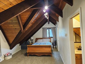 A bedroom with a wooden ceiling, a bed, a nightstand, a lamp, and a window with curtains.