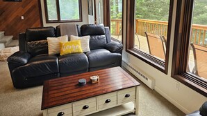 Una sala de estar acogedora con un sofá de cuero negro, una mesa de centro de madera y ventanas amplias que dan a una terraza de madera y árboles.