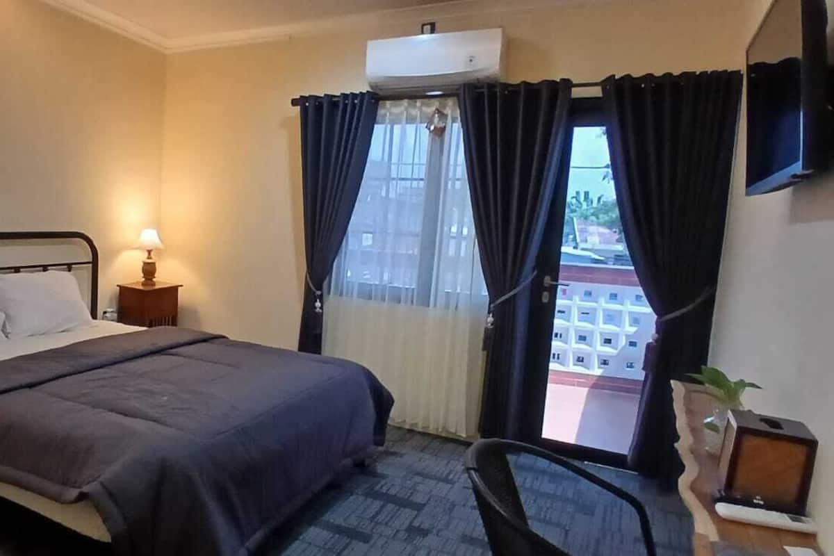 A hotel room with a bed, bedside lamps, a TV, a window with curtains, and a view of buildings outside.