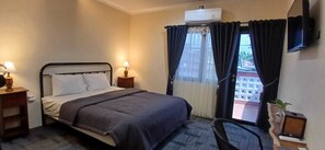 A hotel room with a bed, bedside lamps, a TV, a window with curtains, and a view of buildings outside. - Musafir Guest House Syariah (Surakarta)