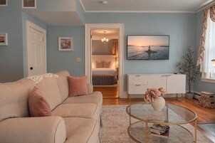 Una sala de estar con un sofá color beige, una mesa de centro de vidrio con libros, un cuadro de un faro y vista al mar a través de una ventana.