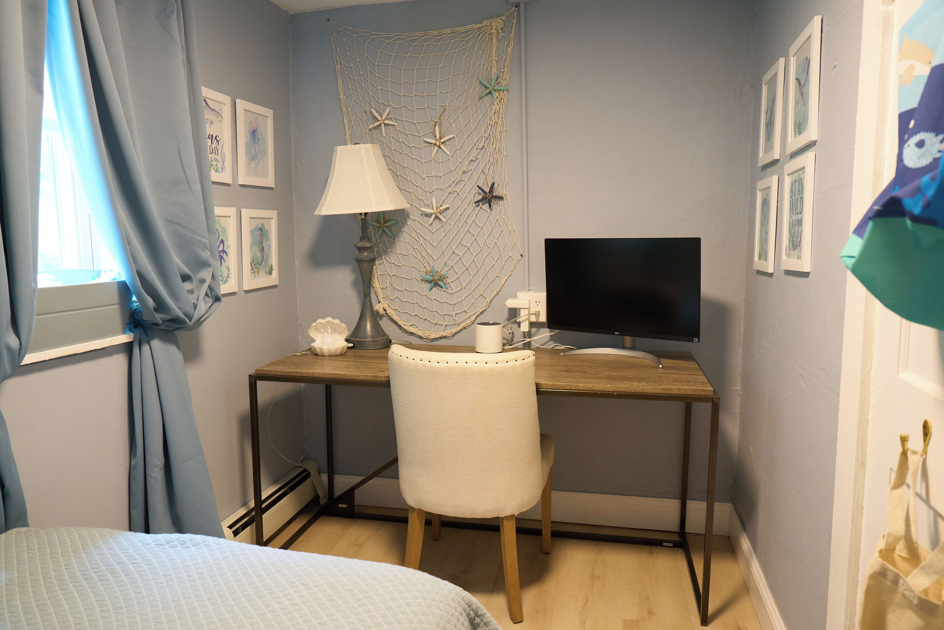 A bedroom with a bed, a desk with a computer, a chair, a lamp, and a wall with framed pictures.