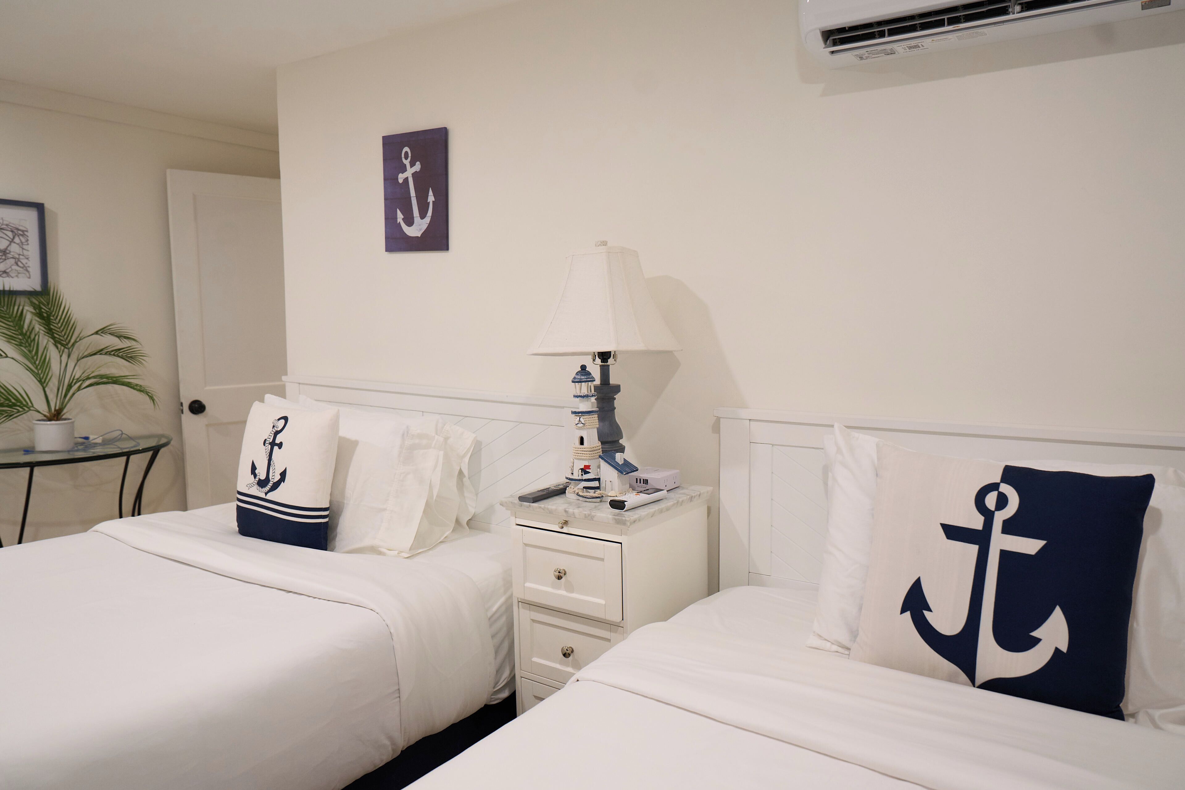A hotel room with two beds, a bedside table with a lamp, a potted plant, and a wall-mounted air conditioner.