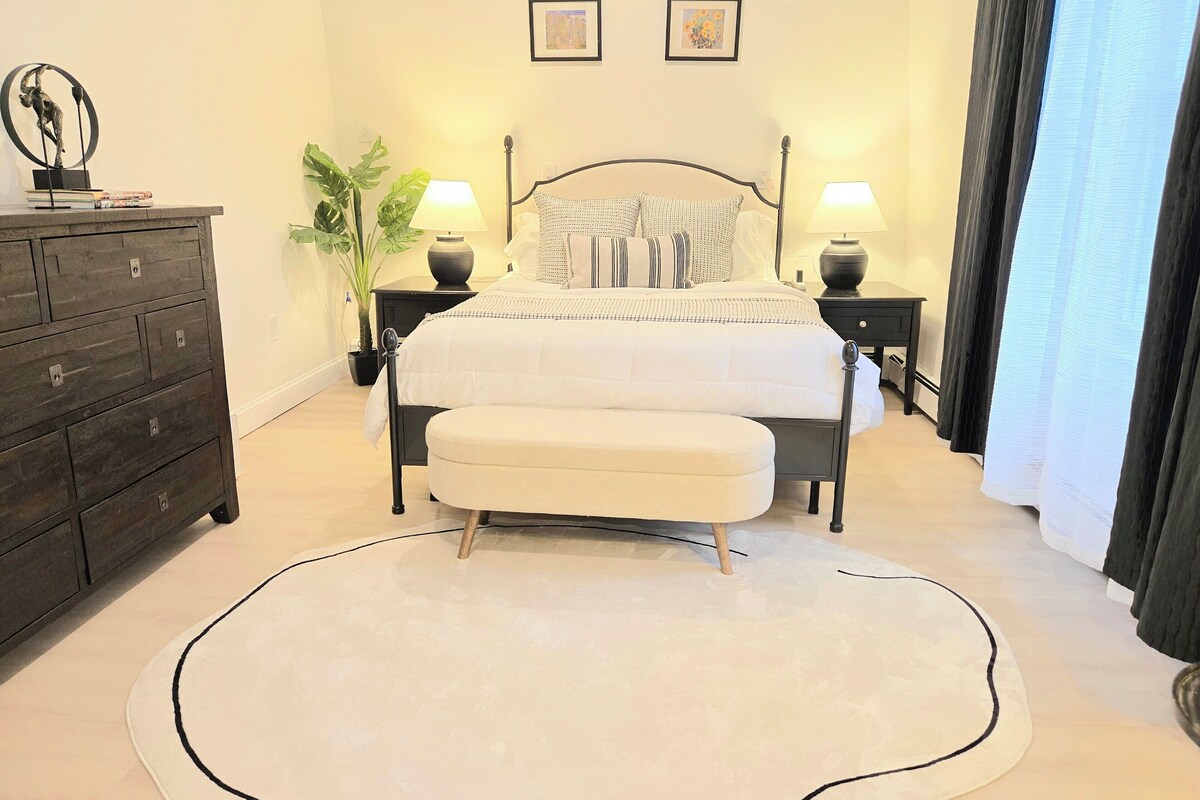Ein Schlafzimmer mit Bett, Nachttischen, einem Schminktisch, einer Topfpflanze und zwei gerahmten Bildern an der Wand.