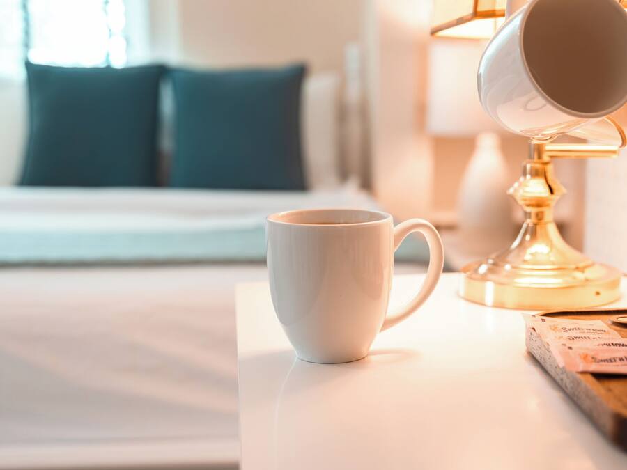 A white cup on a table, a lamp, and a bed with pillows in a hotel room.