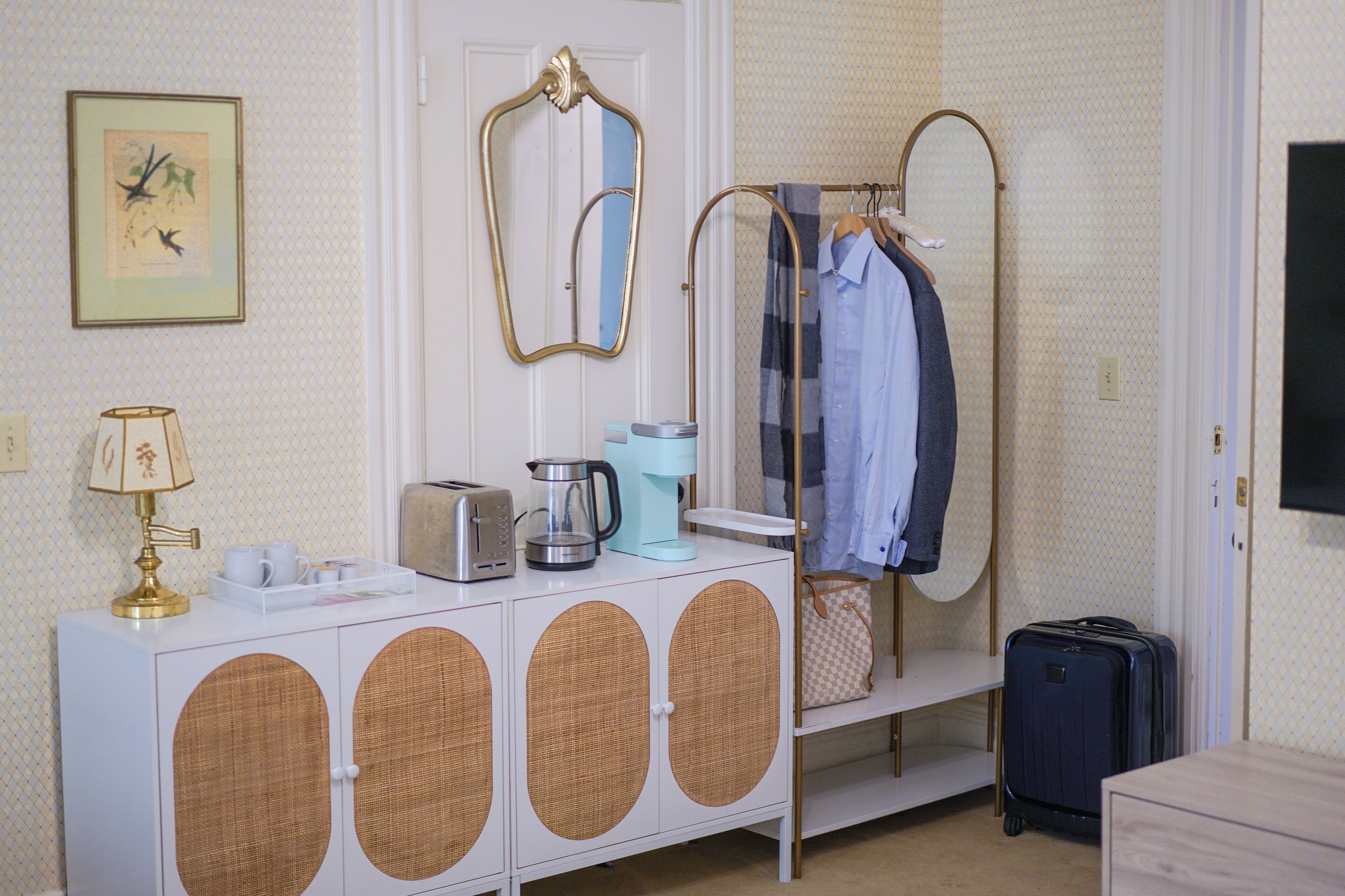 A hotel room with a white console table, a mirror, a coat rack, a suitcase, and a framed artwork on the wall.