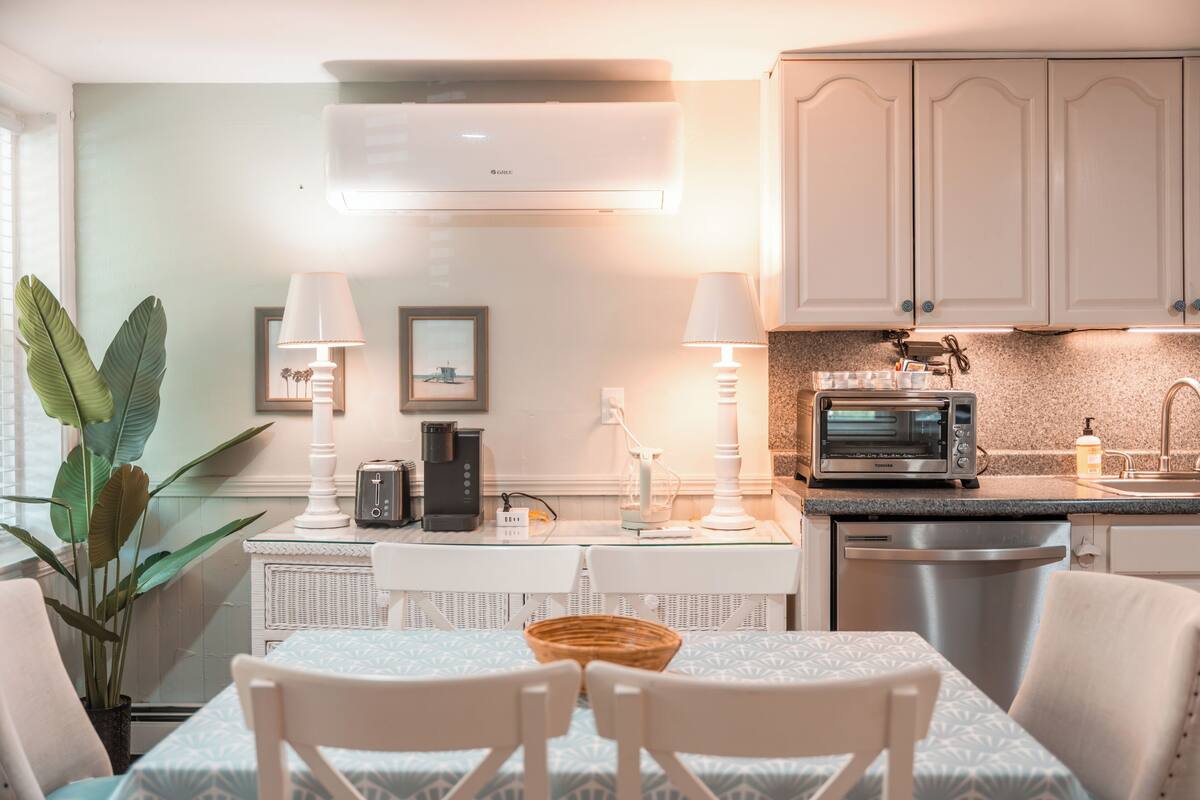 A compact kitchen with a microwave, toaster, and coffee maker on the counter, a dining table with chairs, and a potted plant.