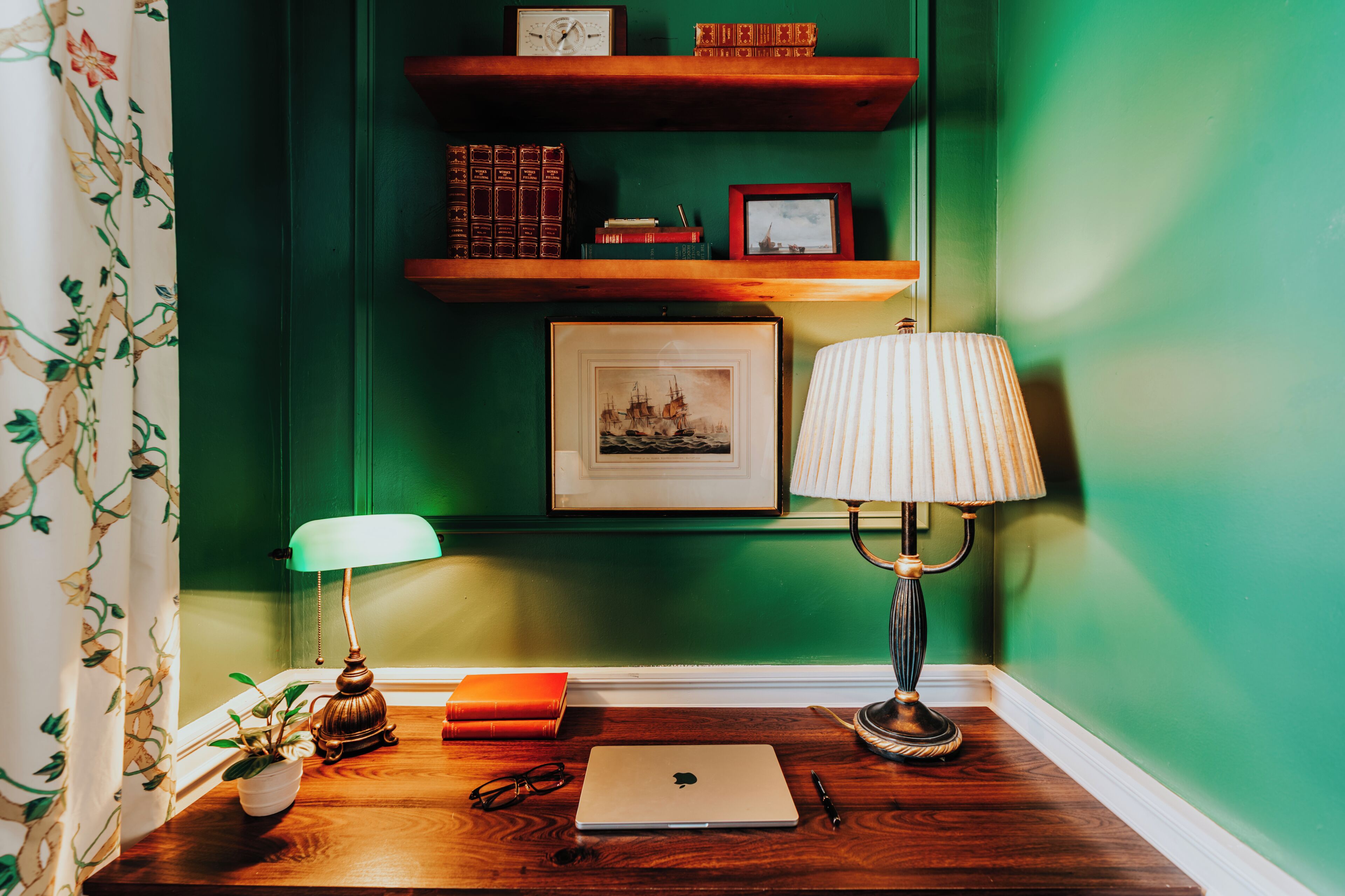 A wooden desk with a laptop, a lamp, a pair of glasses, and a small plant.