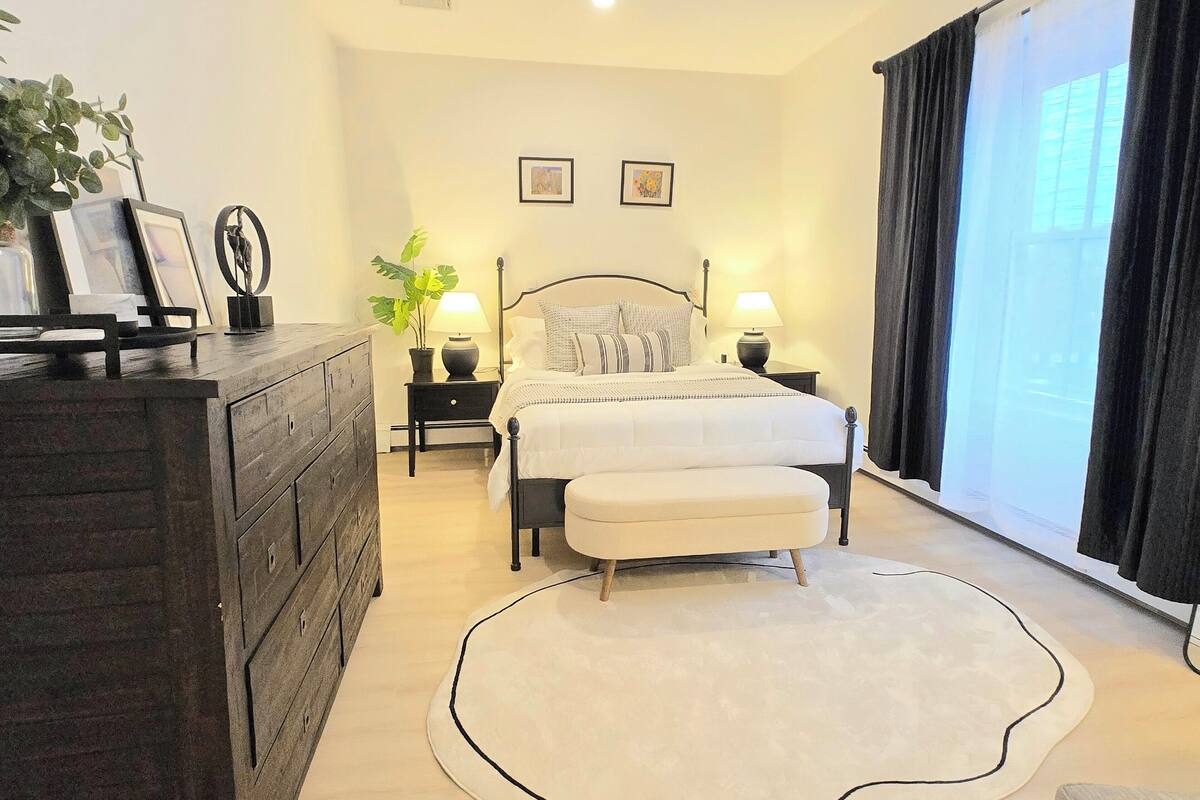 A bedroom with a bed, bedside tables, a dresser, a potted plant, and a large window with curtains.