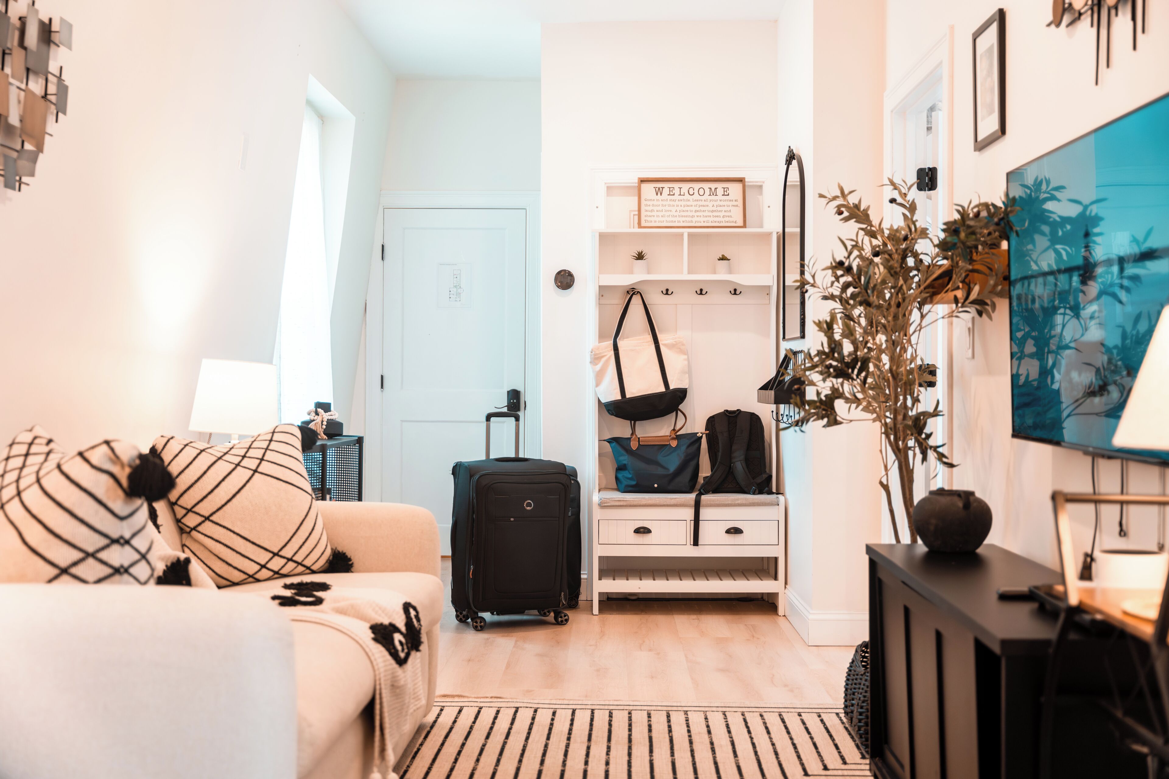 A modern living room with a sofa, a small table, a TV, a wall-mounted shelf with a welcome sign, a plant, and a suitcase.