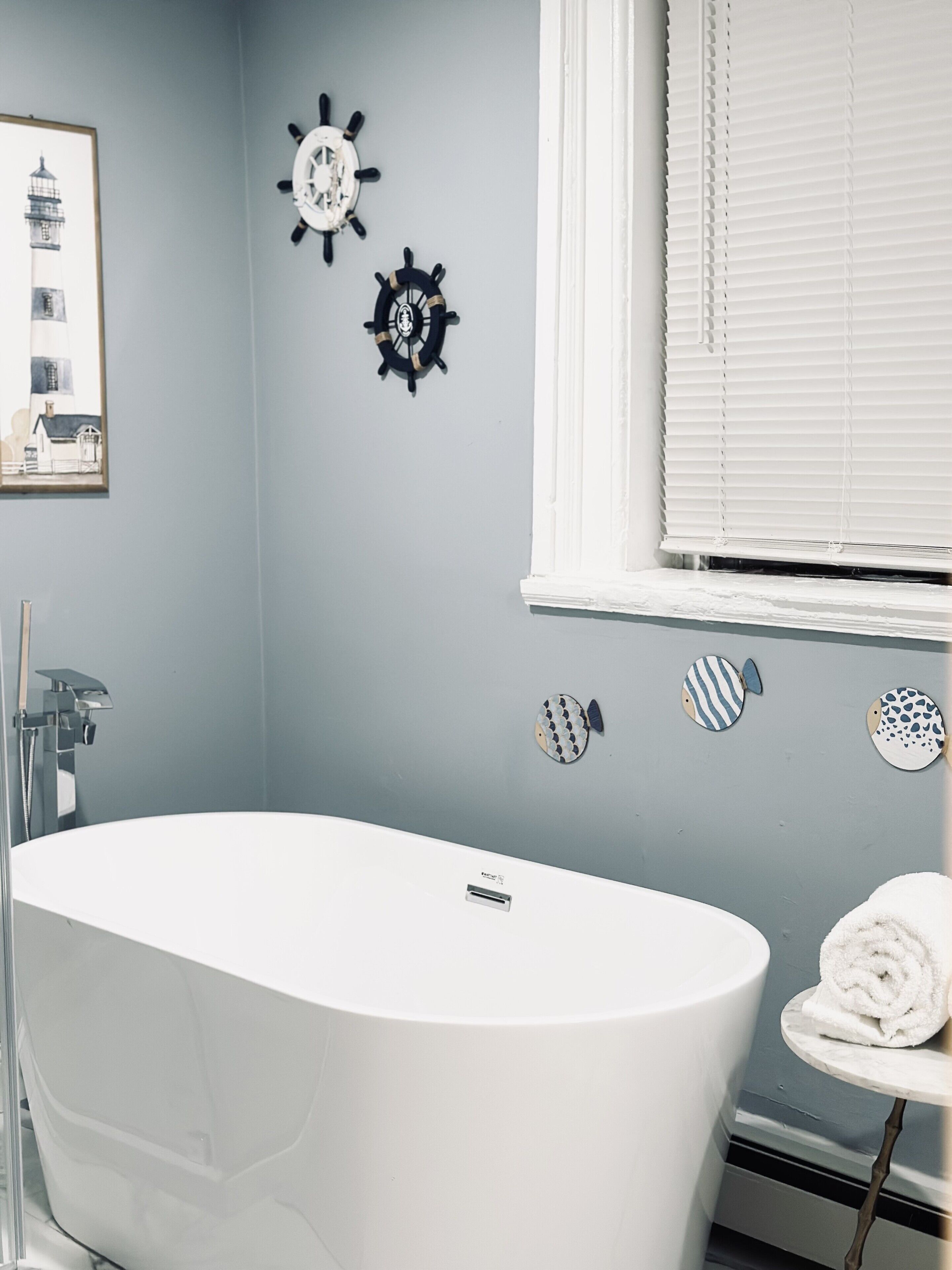 A modern bathroom with a freestanding bathtub, a framed nautical-themed picture, and decorative wall art including ship wheels and fish.