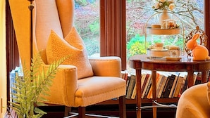 Une pièce comprenant un grand fauteuil, une table avec des livres, une lampe et deux vitraux donnant sur des arbres.