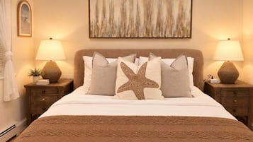 Une chambre à coucher avec un lit, deux tables de chevet avec des lampes, un coussin en forme d’étoile et un tableau abstrait encadré accroché au mur.