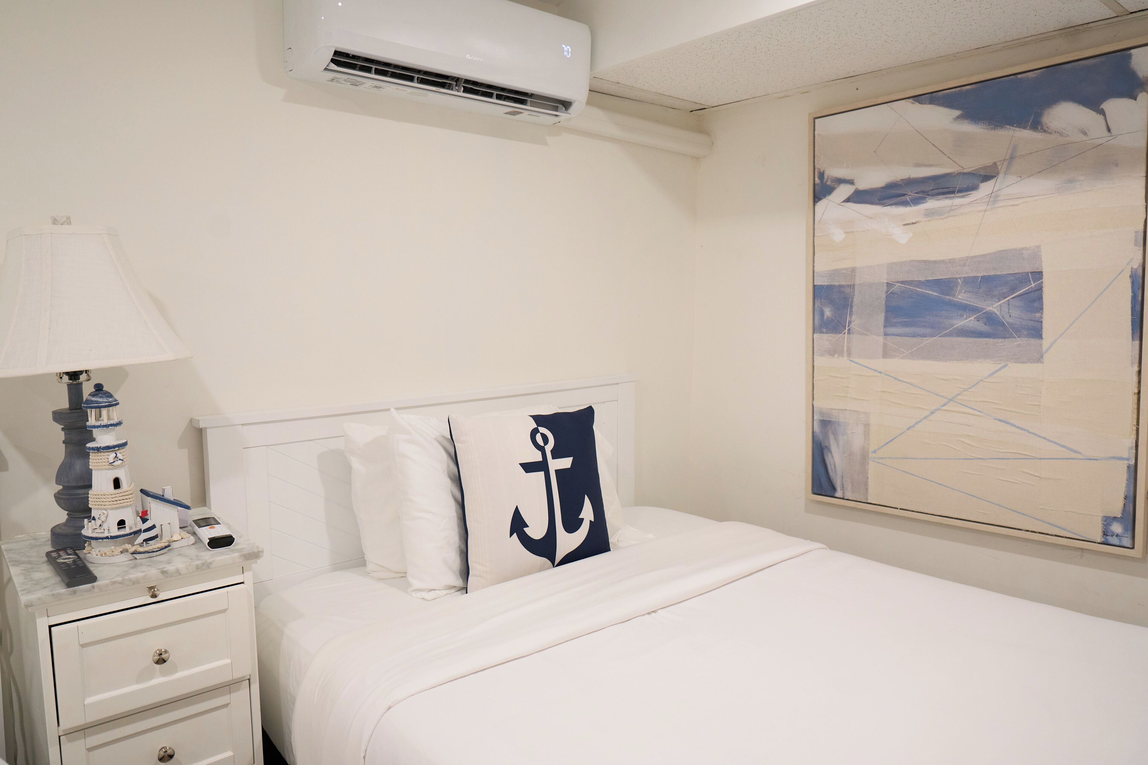 A hotel room with a bed, a nightstand, a lamp, a painting, and an air conditioning unit.