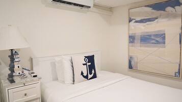 A hotel room with a bed, a nightstand, a lamp, a painting, and an air conditioning unit.