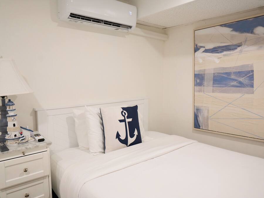 A hotel room with a bed, a nightstand, a lamp, a painting, and an air conditioning unit.