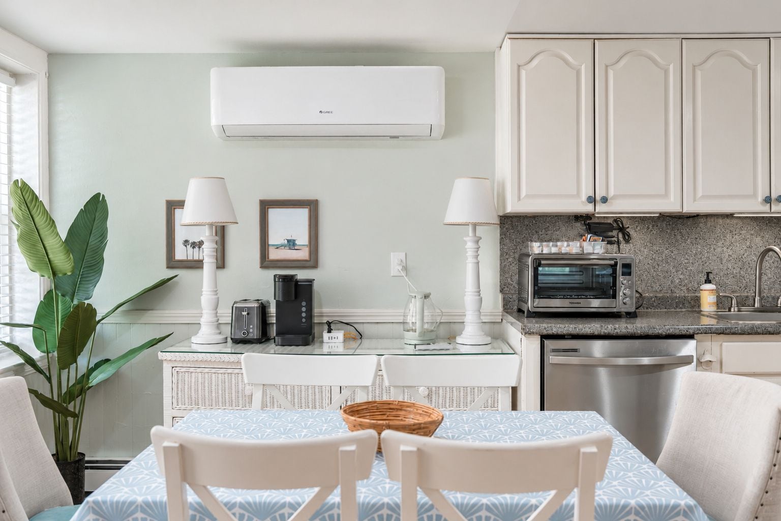 A modern kitchen with white cabinetry, a built-in microwave, and a toaster. There is a dining table with chairs and a large plant.